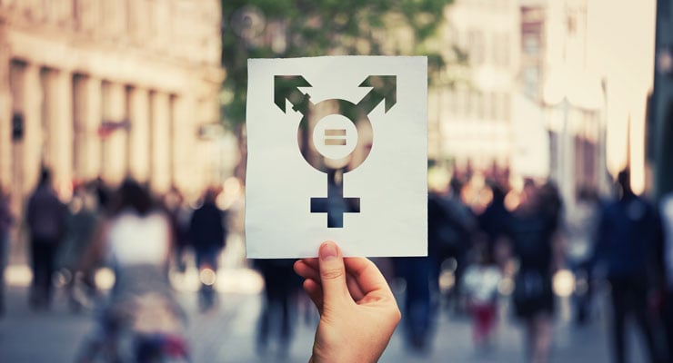 hand holding a paper cutout of a transgender equality symbol in front of a busy sidewalk